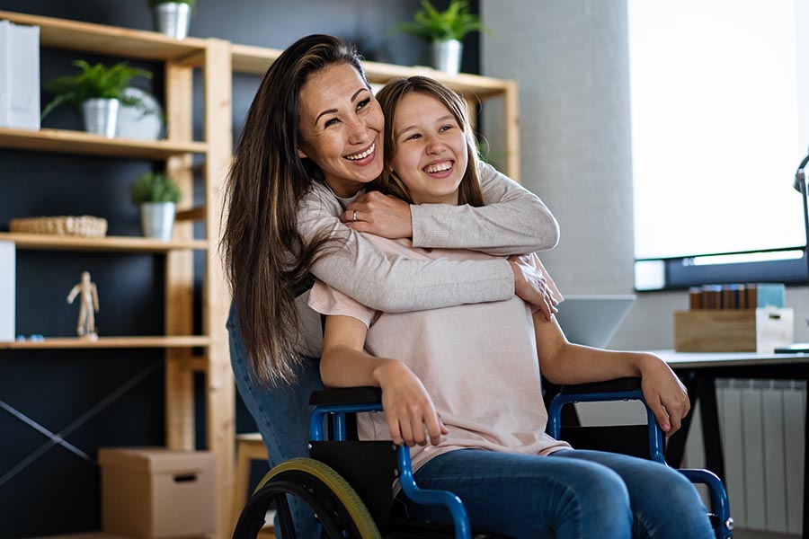 Client Center - Mom Hugs Young Daughter in a Wheelchair, Both Smiling